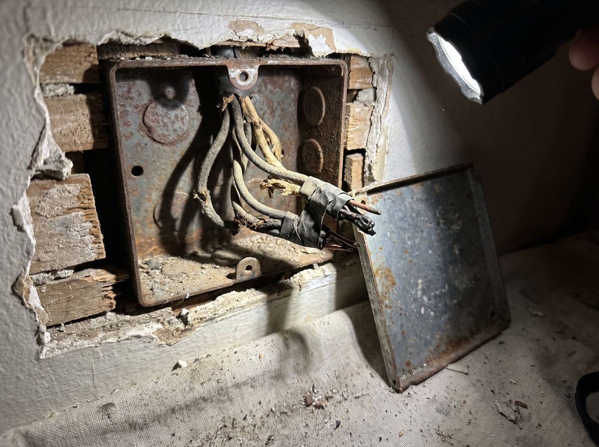 An opened junction box in a wall cavity with brittle cloth-covered conductors and old taped splices.